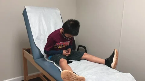 Young boy sits on examination table, waiting for doctor exam, wearing mask. Stock Footage 131400782