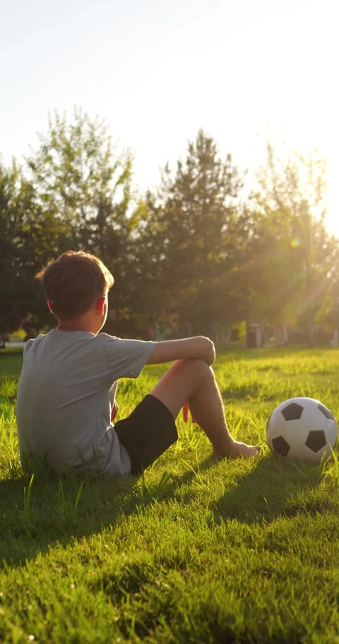 Young boy sits on green grass beside soccer ball in park at sunset. Kid rests Stock Footage 346691355