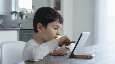 Young boy sits at a table and plays on a tablet in a white kitchen Video stock 128996693