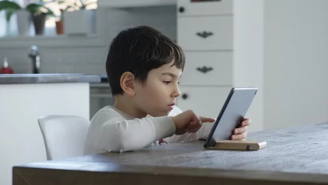 Young boy sits at a table and plays on a tablet in a white kitchen Stock Footage 128996695