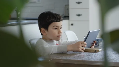 Young boy sits at a table and plays on a tablet in a white kitchen Video stock 128996756