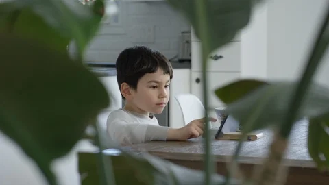 Young boy sits at a table and plays on a tablet in a white kitchen Video stock 128996763