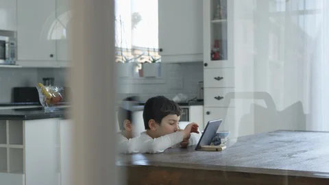Young boy sits at a table and plays on a tablet in a white kitchen Video stock 128996810
