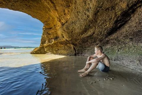 Young boy sitting in a cave looking at the ocean on the beach Foto stock