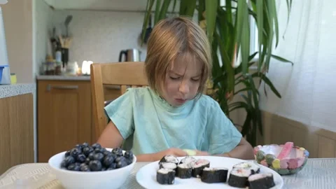 Young boy sitting at the kitchen table eating cucumber with a plate of sushi Stock Footage 318220317