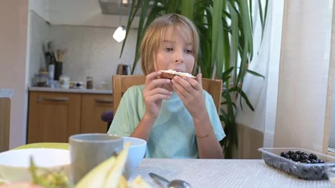 Young boy sitting at the kitchen table eating a sandwich with cream cheese, with Stock Footage 318224603