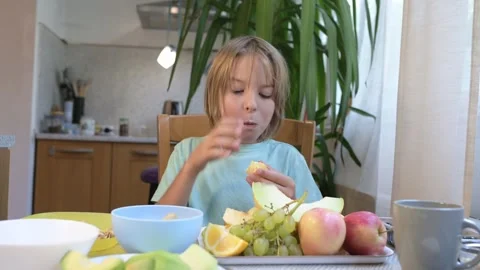 Young boy sitting at the kitchen table eating fresh fruits including orange Stock Footage 318225379