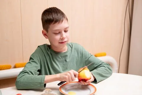 Young boy sitting at the kitchen table and using a knife to remove the skin.. Stock Photos
