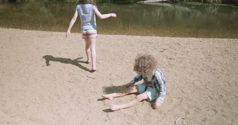 Young boy sitting on a river beach covers himself with sand, young girl walks by 库存影片 99358149