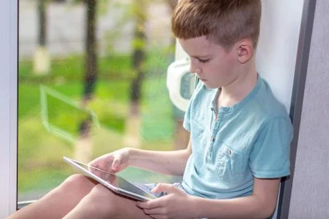 Young boy sitting on windowsill with tablet and play the games. Stay at home Stock Photos