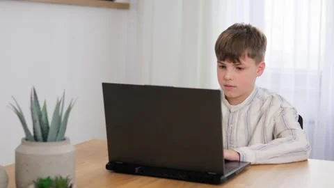 A young boy is sitting, working thoughtfully at his laptop and looking at the Stock Footage 272436472