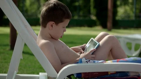 Young Boy With Smartphone or Tablet Sitting in neatr the Pool on a chair. Play Stock Footage 80685207