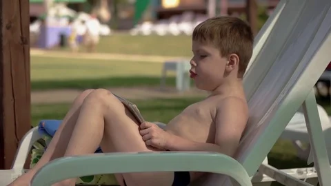 Young Boy With Smartphone or Tablet Sitting in neatr the Pool on a chair. Play Stock Footage 80685360