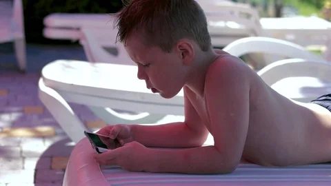 Young Boy With Smartphone or Tablet Sitting in neatr the Pool on a chair. Play Stock Footage