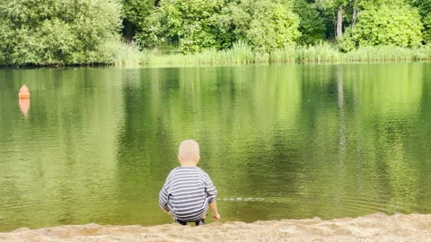 A young boy squats and touches the water at Kolonistsky Pond Beach in Pushkin Video stock 344987316
