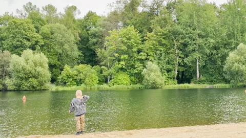 A young boy squats playfully on the sandy shore by the pond in Pushkin Video stock 344986628