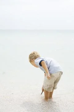 Young boy standing ankle deep in water at the beach, bending over, looking down 写真素材