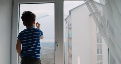 A young boy is standing in front of a window while looking outside Stock Footage 306673555