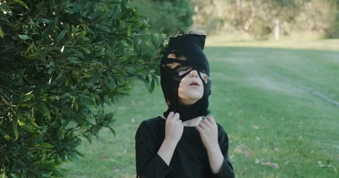 A young boy standing under a tree in a park pulls on a Halloween ninja mask  Stock-Footage 115787319