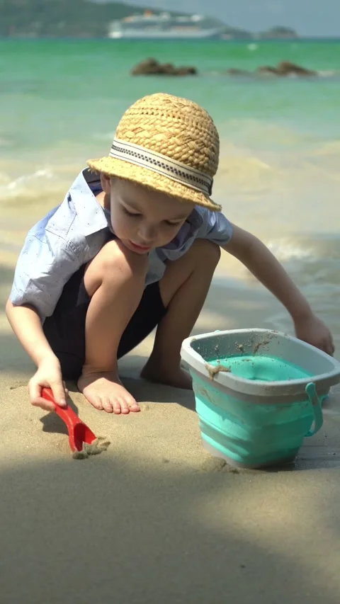 Young boy in a straw hat crouches on the sand, filling a turquoise bucket Vídeo Stock 347101899