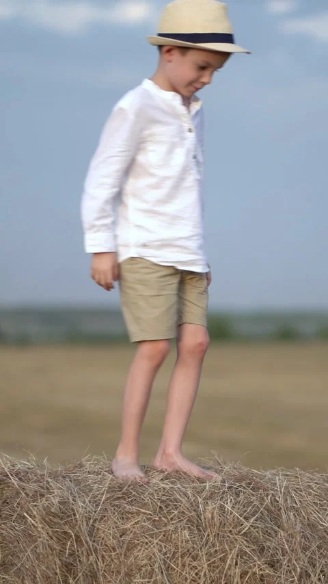 Young boy in straw hat stands and walks barefoot on a hay bale in a dry rural  Stock Footage 341530386