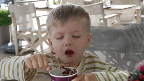 Young boy in a striped shirt eats chocolate ice cream with a spoon at a cafe. He Stock Footage 302543024