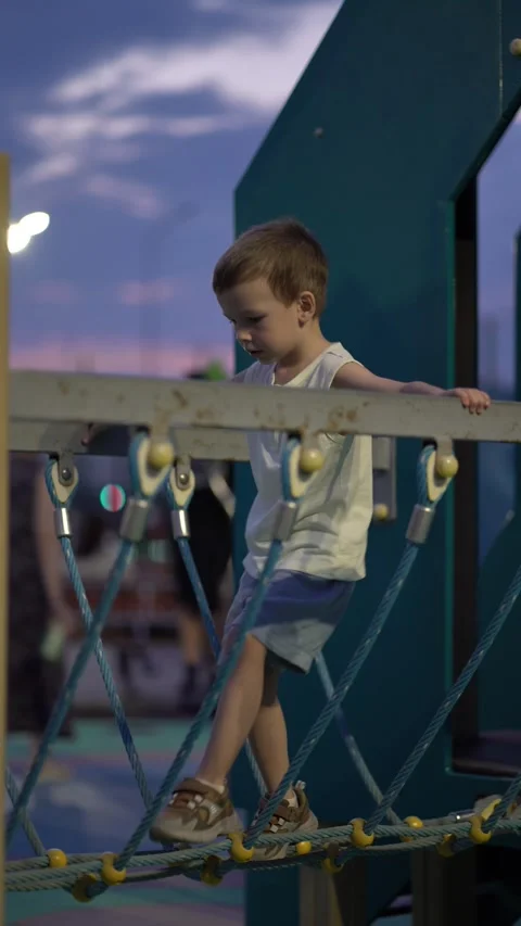 Young boy thoughtfully balances on a rope bridge at the playground during dusk Stock Footage 341410050