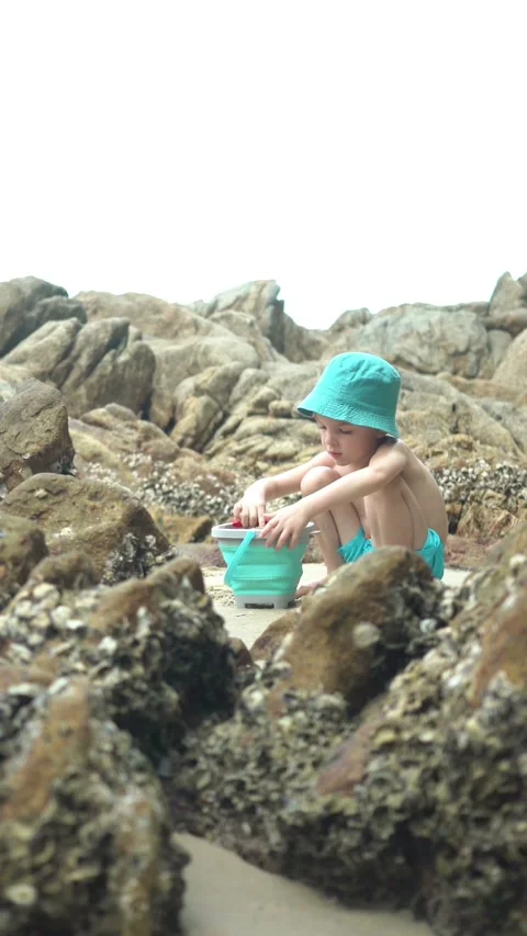 A young boy in a turquoise hat digs in the sand with a blue shovel beside 스톡 동영상 347101817