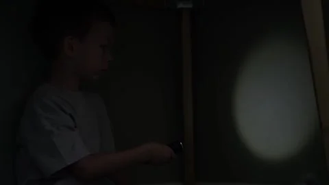 Young boy under table in dark room shines flashlight on wall creating circular 動画素材 346305714