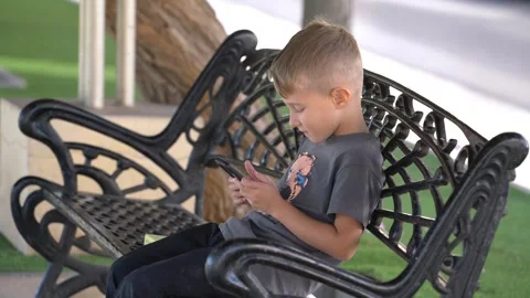 Young boy using smartphone while sitting on park bench outdoors 스톡 동영상 323566169