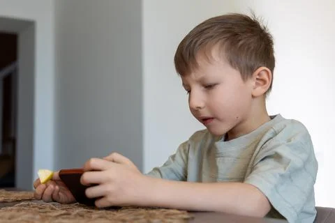 Young boy using smartphone while eating fruit. A young boy sits at a table Stock Photos