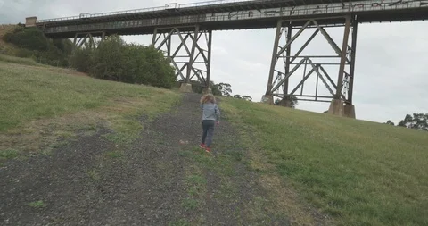 Young boy walking and running up a hill in a park near a railway trestle bridge 库存影片 95291206