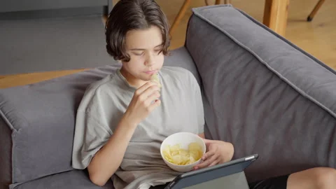 Young boy watching streaming service on a tablet and eating chips at home Stock Footage 315560606