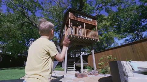 Young boy waves to his sisters in the treehouse above in their backyard Stock Footage 245766228