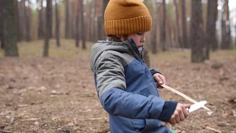 A young boy wearing a beanie skillfully whittles a stick with a knife in a Video stock 291168328