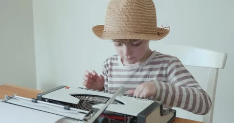 Young boy wearing a hat is learning to type on an old portable typewriter 库存影片 127966628