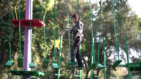 A young boy wearing a safety harness carefully walks and balances across hanging Stock Footage 345285683