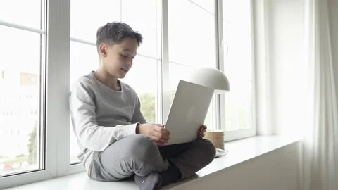 Young boy working on a laptop computer while relaxing on the windowsill at home Stock Footage 142924947