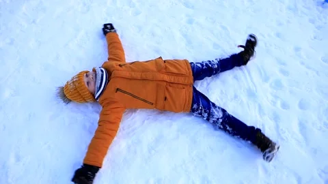 A young boy in a yellow jacket lies in the snow and makes a snow angel. Stock-Footage 160708108