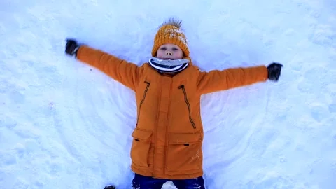 A young boy in a yellow jacket lies in the snow and makes a snow angel. Video stock 160788436