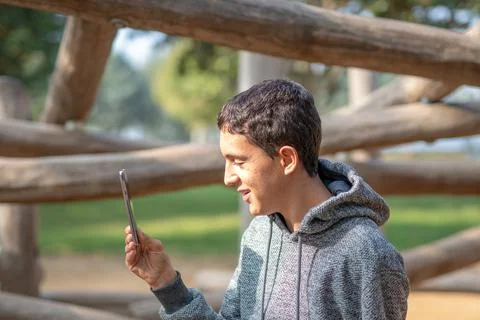Young brown-haired boy looking at his cell phone in a park wearing a gray hoo Stock Photos