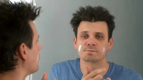 Young brunet man ia applying moisturizing cream on his face looking at mirror. Stock Footage 168224591