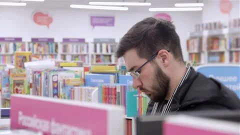 Young brunette black-haired man with glasses looking books on the bookshelves in Stock Footage 88801412