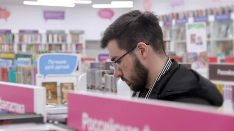 Young brunette black-haired man with glasses looking books on the bookshelves in Stock Footage 88801607