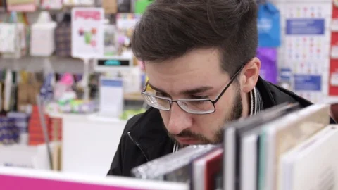 Young brunette black-haired man with glasses looking books on the bookshelves in Stock Footage 88801675