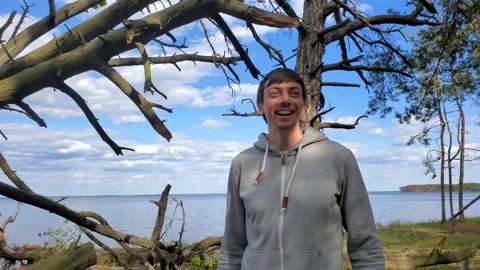 Young brunette man is standing in a pine forest by the lake and talking Видео 130575780