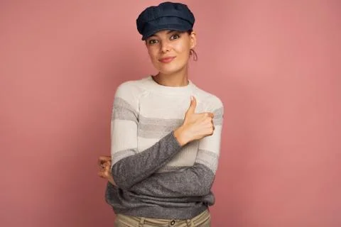 Young brunette in a pullover and blue cap smiles at the camera with her thumb up Stock Photos