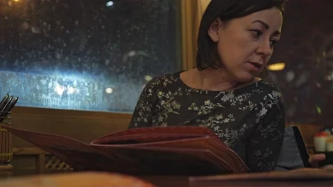 A young brunette is sitting at a table in a restaurant and choosing dishes from Stock Footage 170565949