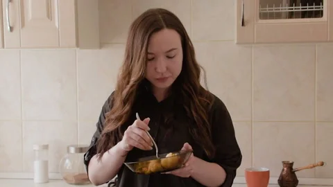 A young brunette is standing in the kitchen eating steamed hot vegetables. Stock Footage 107193423