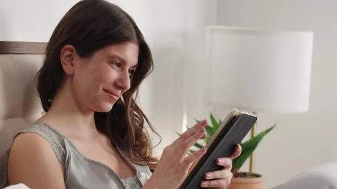 A young brunette uses a digital tablet while sitting on a bed by the window. A Stock Footage 245683339